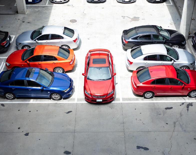 Luftfoto af en parkeringsplads med flere rækker af parkerede biler i forskellige farver, markeret med hvide linjer og pile på asfalten.